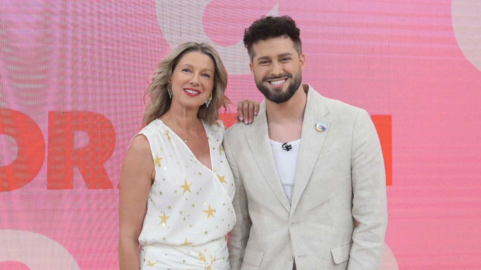 Anne Igartiburu y Javi Hoyos sonríen a cámara. Ella viste blanco con estrellas doradas; él, americana beige y pin. Fondo rosa con letras rojas 'OR'.