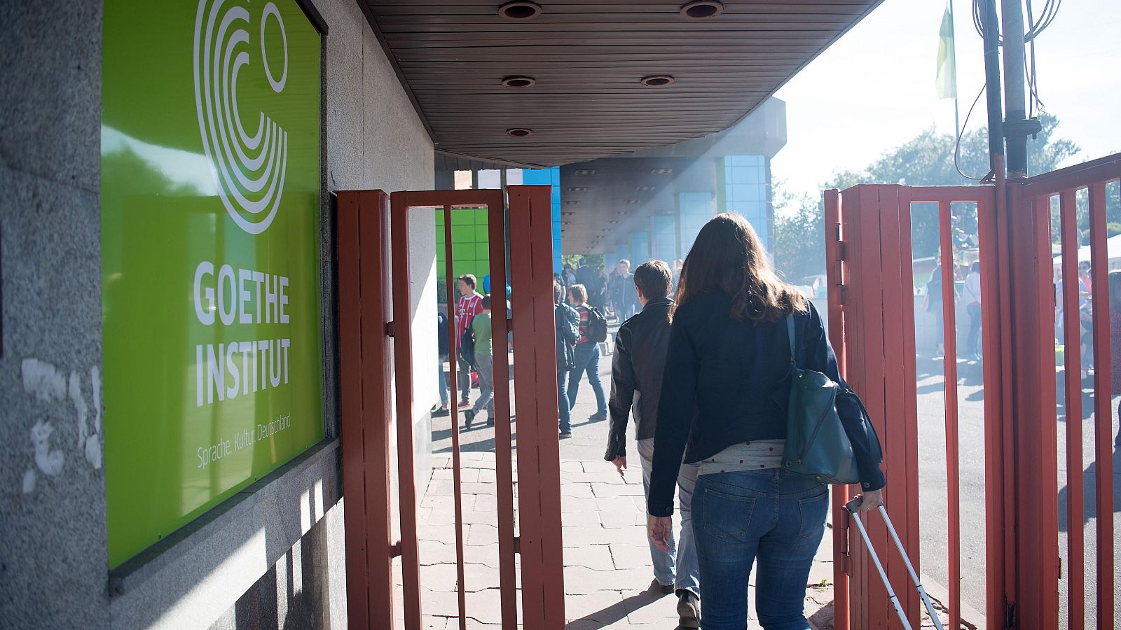 Entrada del Goethe-Institut en Moscú con cartel verde y mujer de espaldas entrando. Se aprecian personas en la distancia bajo luz solar.