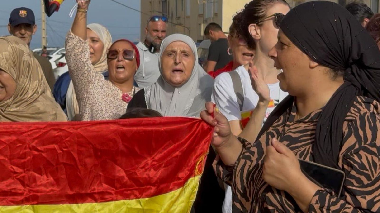 Manifestación en Ceuta con mujeres expresando sus demandas, sosteniendo una bandera de España. Se aprecian edificios urbanos y vehículos al fondo.
