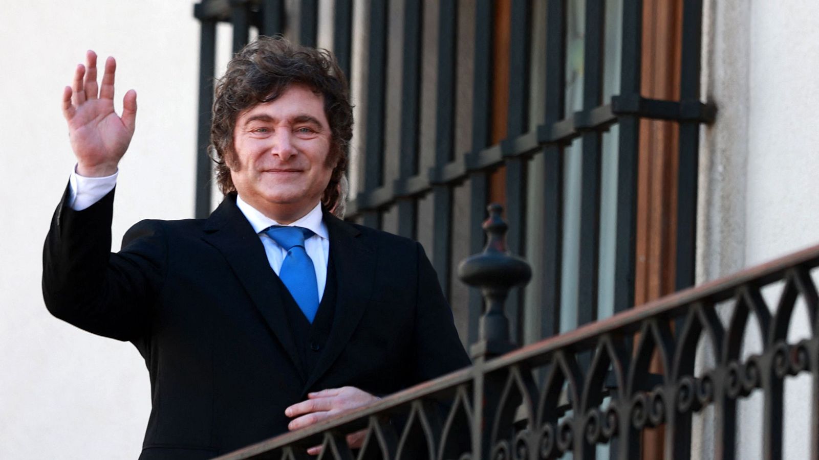 Javier Milei, Presidente de Argentina, sonríe y saluda con la mano derecha levantada. Viste traje negro, camisa blanca y corbata azul. Se encuentra en un balcón.