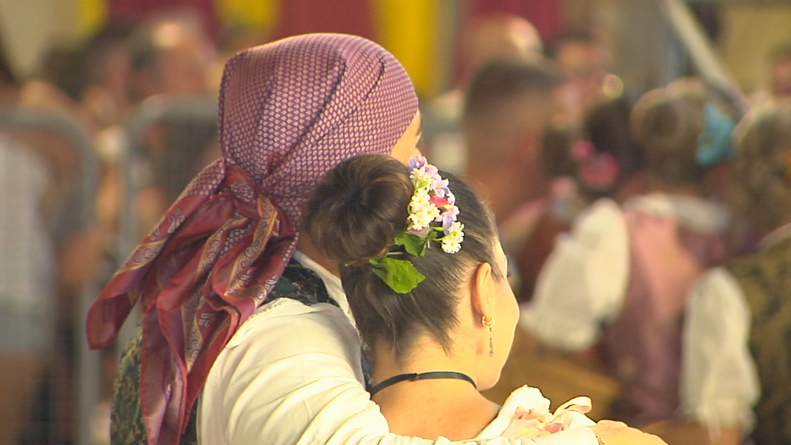 Dos mujeres con atuendos tradicionales asisten a una celebración festiva. Una lleva un moño adornado con flores y la otra un pañuelo morado y granate.