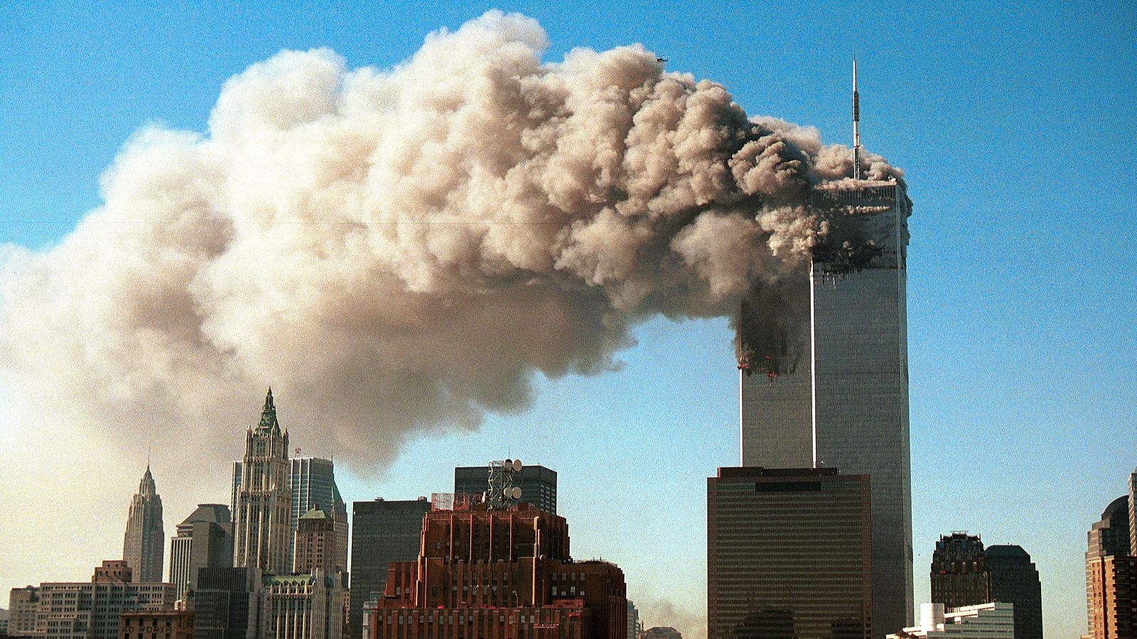 Vista de las Torres Gemelas del World Trade Center en llamas y cubiertas de humo denso, con el skyline de Manhattan al fondo bajo un cielo azul.