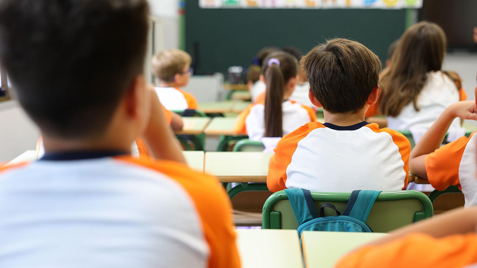 Vista posterior de un aula con niños uniformados (camiseta blanca y naranja) sentados en pupitres. Al fondo, una pizarra oscura. Ambiente educativo.