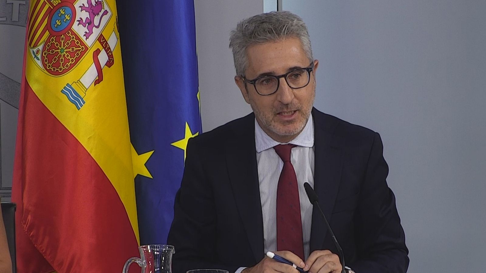Carlos Cuerpo, Ministro de Economía, Comercio y Empresa, habla en una rueda de prensa, con las banderas de España y la UE de fondo.