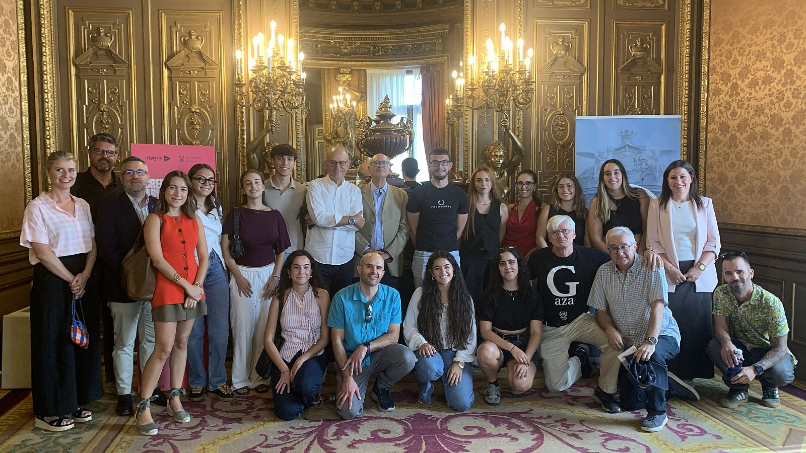 Veintitrés personas posan en un salón ornamentado. Destacan un hombre con camiseta de Fred Perry y otro con una camiseta que dice "Gaza". Al fondo, carteles de RTVE y un estandarte azul.