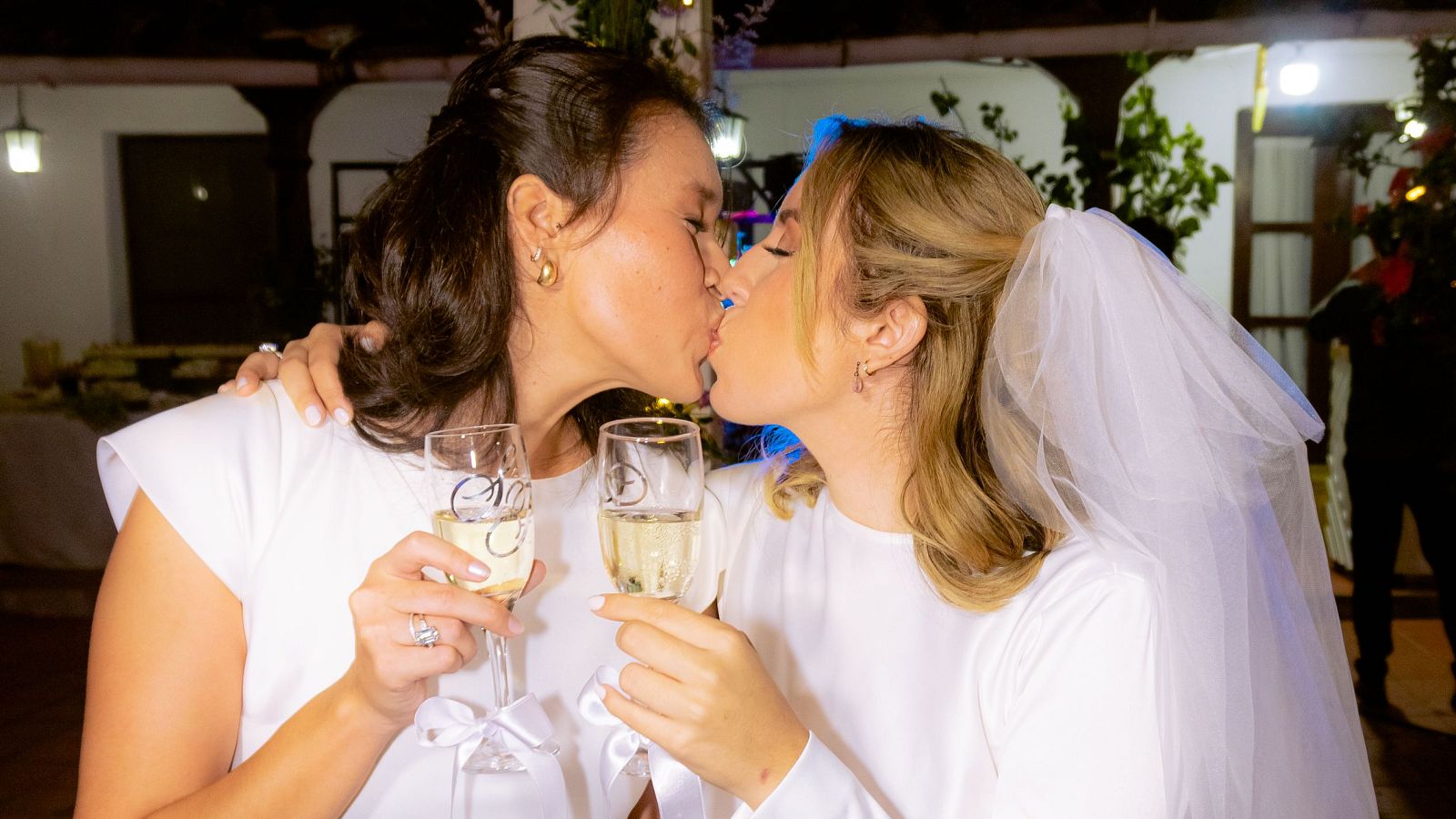 Fabiana y Scarlett, recién casadas, comparten un beso tierno y emotivo, con copas de champán en mano y anillos brillando, en un entorno nocturno decorado con luces.