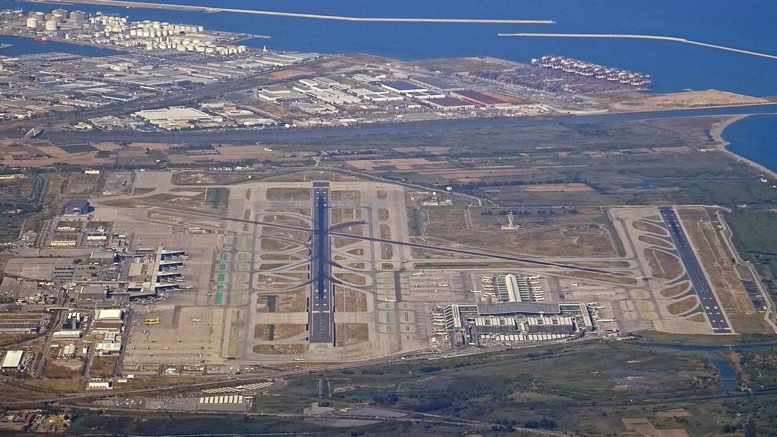 Vista aèria de l'Aeroport de Barcelona-El Prat, mostrant pistes, terminals T1 i T2 amb aeronaus, torre de control, el Port de Barcelona i zones industrials.