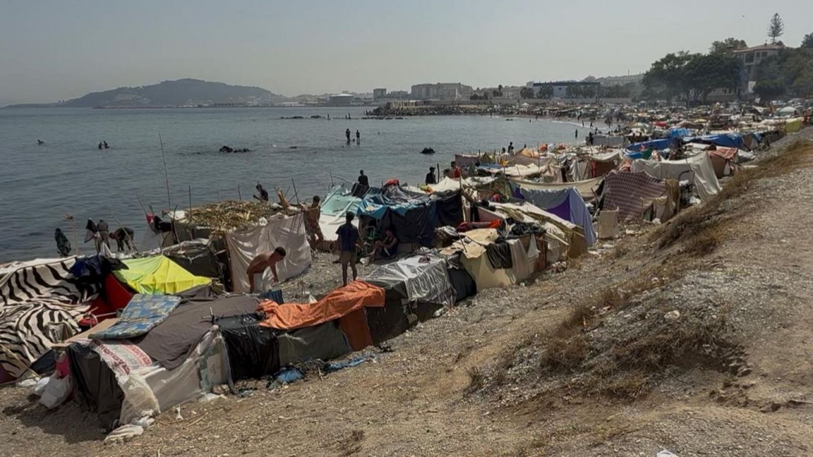 Vista panorámica de la Playa del Trampolín en Ceuta, transformada en un campamento de migrantes con refugios improvisados y numerosas personas ocupando la costa bajo un cielo despejado.