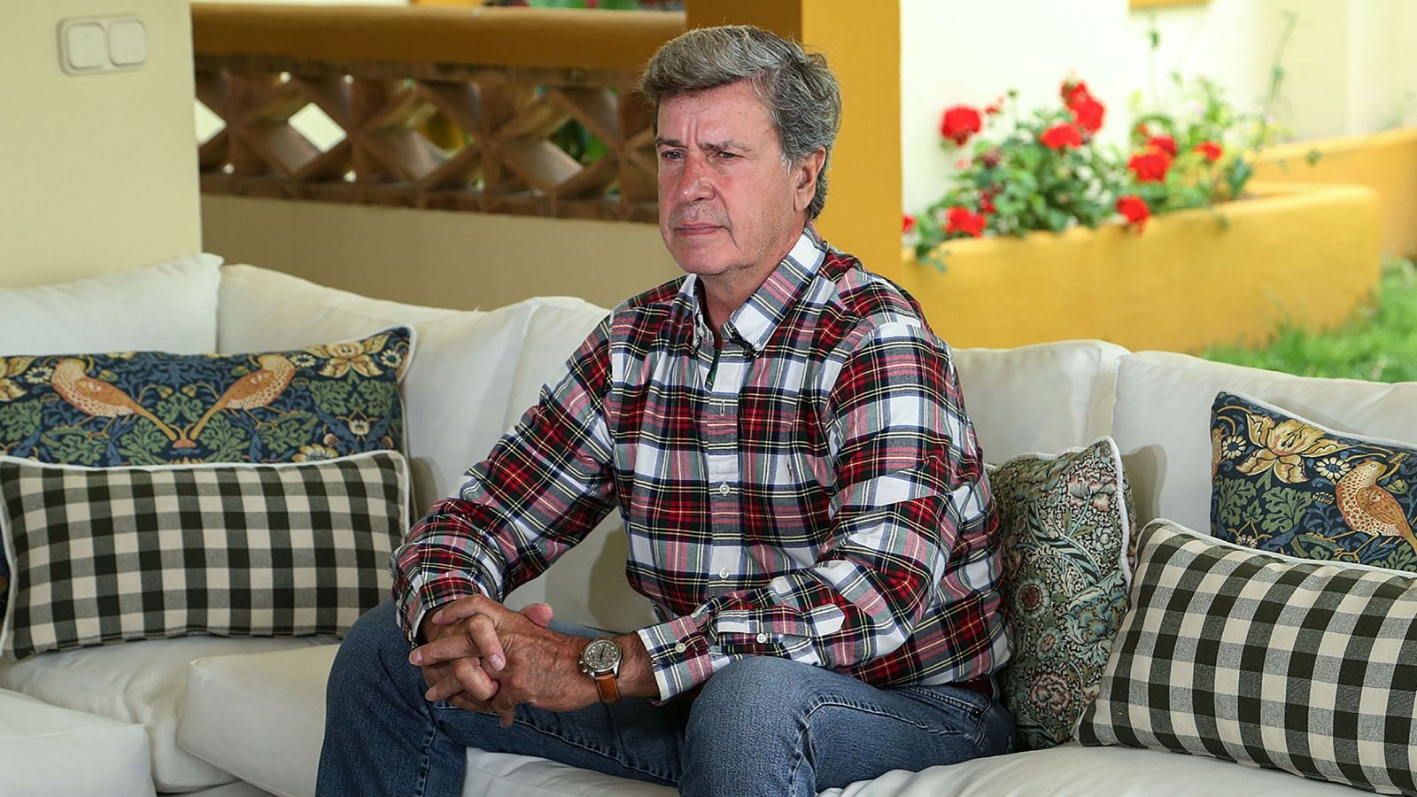 Cayetano Martínez de Irujo, con camisa de cuadros y vaqueros, posa en un sofá con cojines. Detrás, una pared beige y un jardín con flores rojas.
