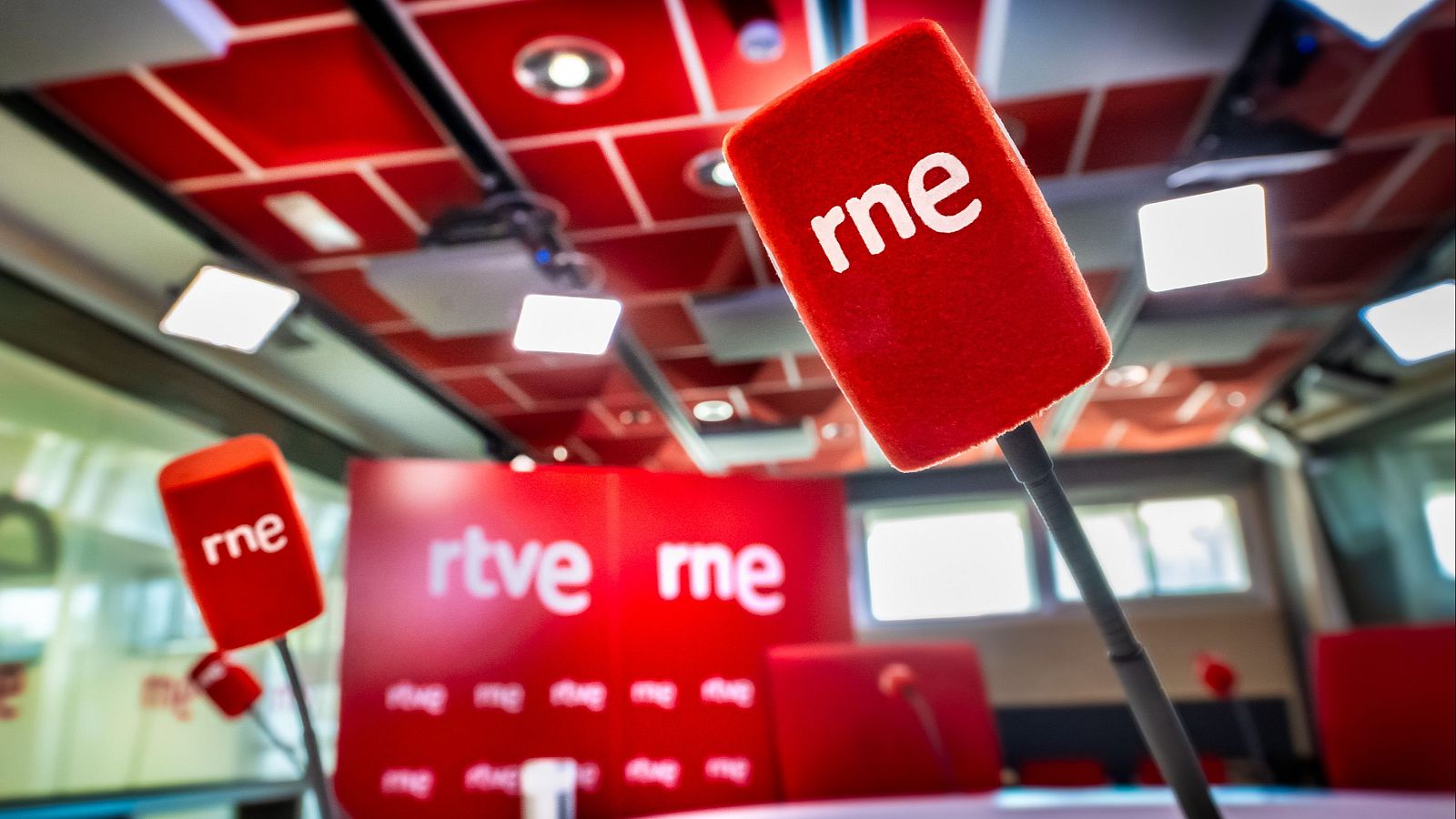 Estudio de radio con micrófonos "rne" y panel de fondo "rtve" y "rne" en rojo y blanco. Luces empotradas en techo rojo.
