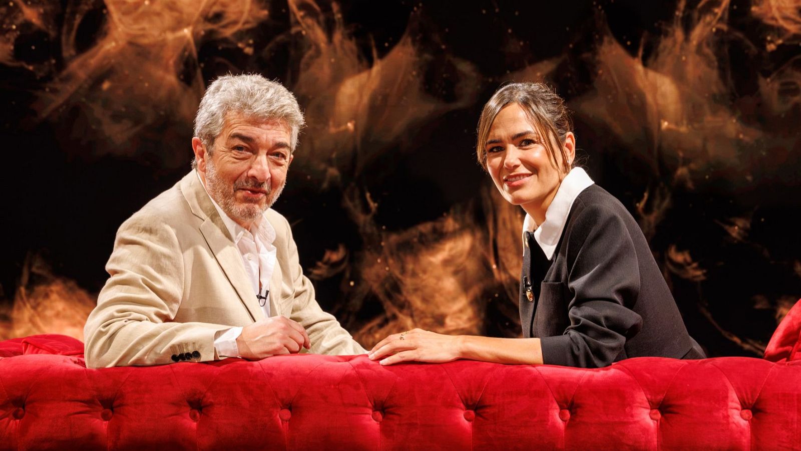 Ricardo Darín y Elena Sánchez, sentados en un sofá rojo, sonríen a la cámara. Darín viste chaqueta beige y Elena chaqueta oscura con camisa blanca. Fondo abstracto cálido.