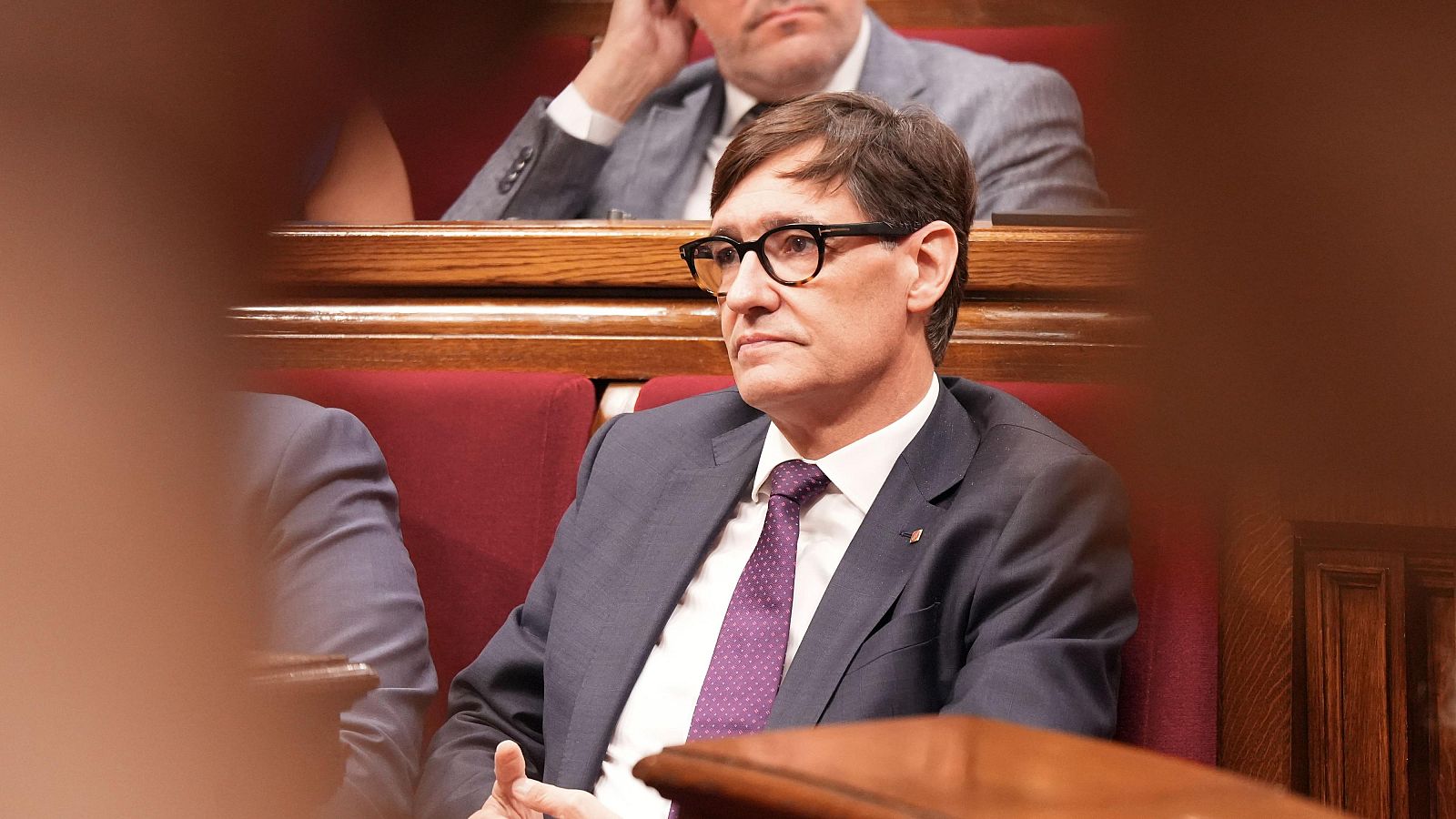 Primer pla de Salvador Illa, polític espanyol, assegut en un hemicicle. Porta ulleres, vestit fosc i corbata morada. Entorn parlamentari amb seients vermells i fusta fosca al fons.