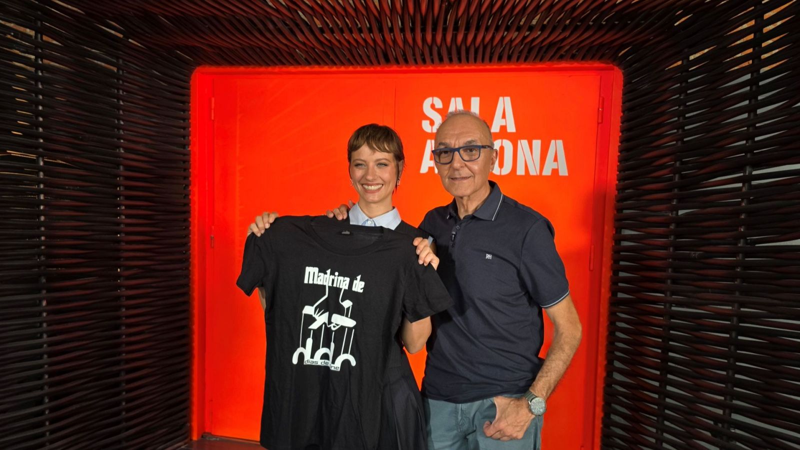 Michelle Jenner y Gerardo Sánchez sonríen. Ella sostiene una camiseta de "Madrina de Días de Cine" con marionetas. Él, con gafas, apoya su brazo en su hombro. Fondo naranja con "SALA MADRONA" y mimbre.