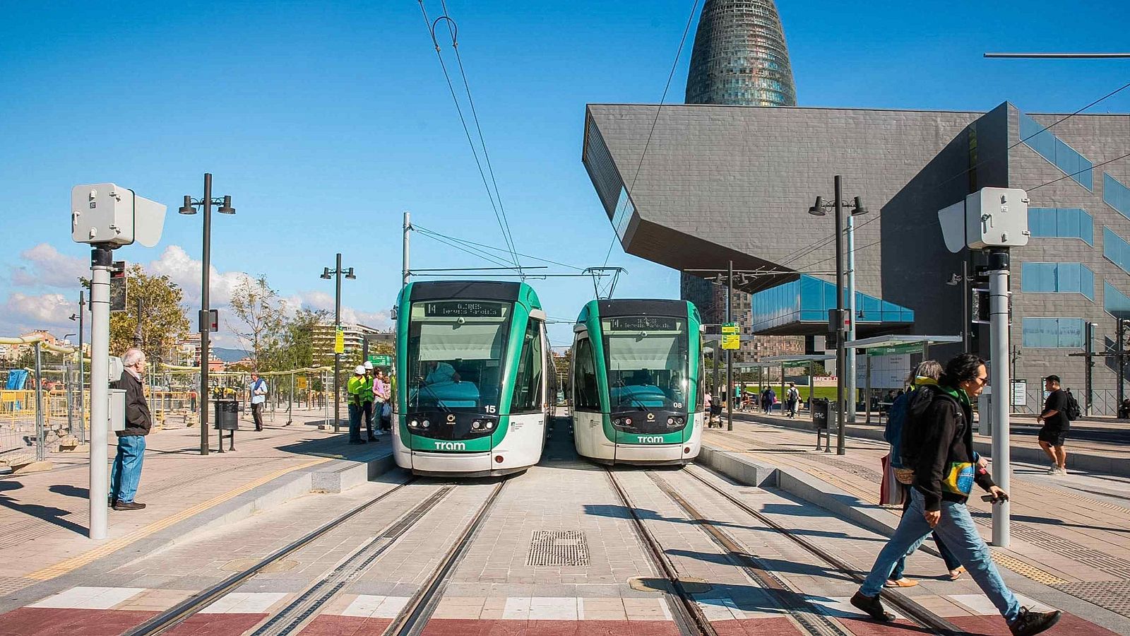 Dos tranvías verdes y blancos con la palabra "TRAM" en el frontal están detenidos en la estación de Glòries, Barcelona, bajo un cielo azul. Personas y la Torre Glòries completan la escena urbana.