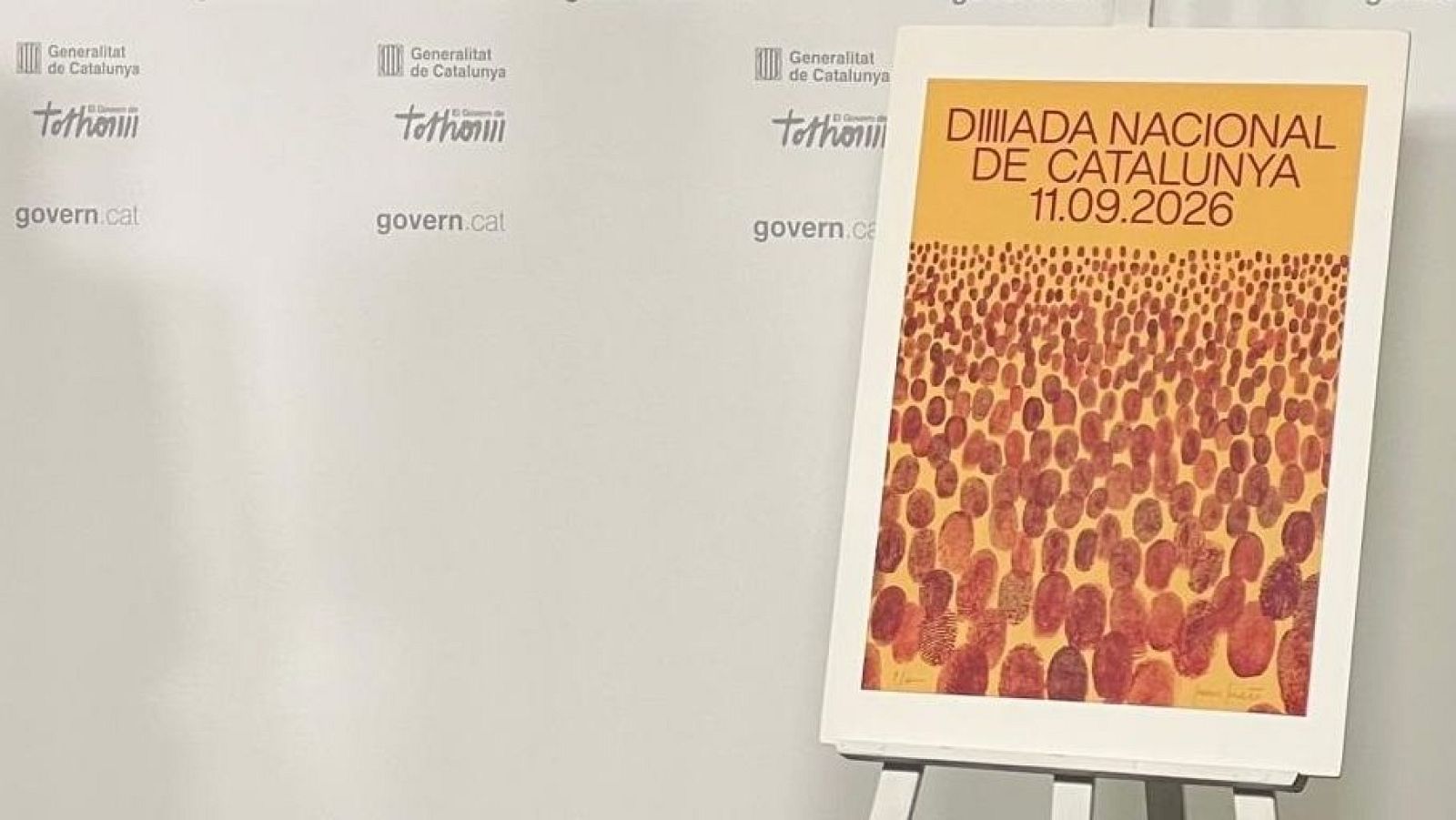 Cartell de la Diada Nacional de Catalunya 11.09.2026 amb un disseny d'empremtes dactilars en vermell i marró sobre fons groc, recolzat en un cavallet davant una paret amb logotips de la Generalitat.