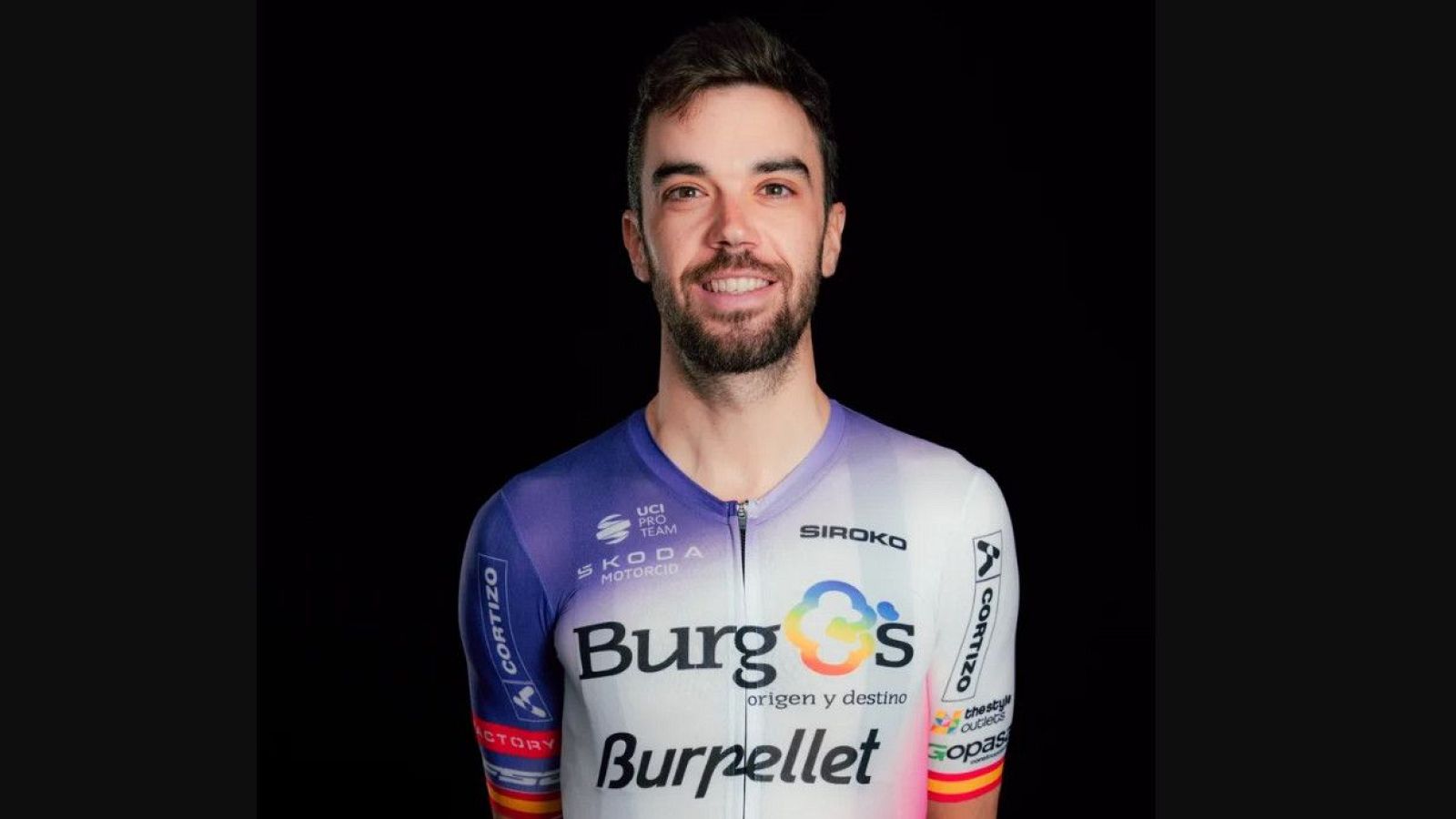 Retrato de Jesús Herrada, ciclista profesional, luciendo un maillot blanco y morado/azul con "Burgos" y "SIROKO" visibles. Fondo negro.