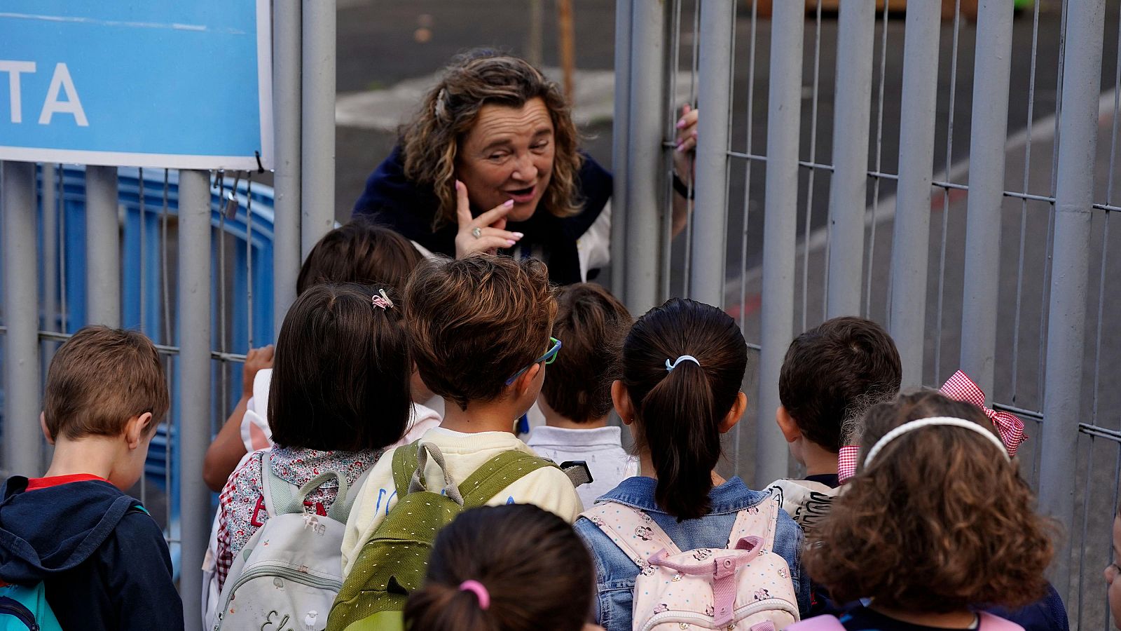 Profesora de pelo rizado interactúa con escolares de Primaria a través de una verja gris en la puerta de un colegio. Los niños la observan atentamente.