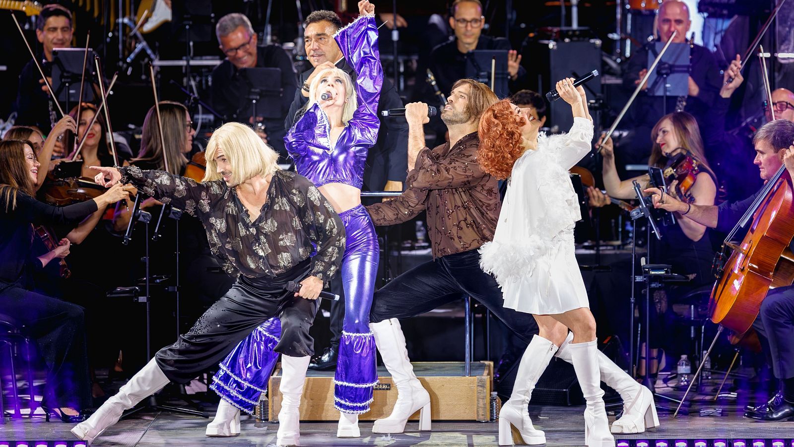 Cuatro artistas imitan a ABBA con trajes brillantes y botas de plataforma, acompañados por una orquesta en un concierto en vivo.
