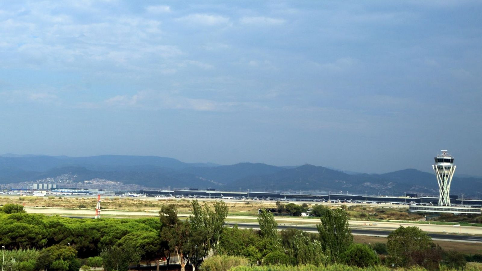 Vista panorámica del Aeropuerto del Prat mostrando vegetación, pistas de aterrizaje, torre de control y montañas a lo lejos bajo un cielo parcialmente nublado.