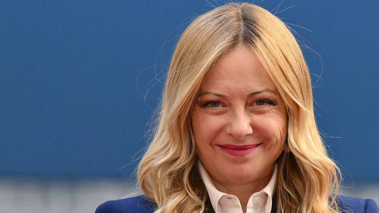 Giorgia Meloni, primera ministra de Italia, sonríe con arrugas de expresión. Cabello rubio ondulado, camisa clara y blazer azul. Fondo azul liso.