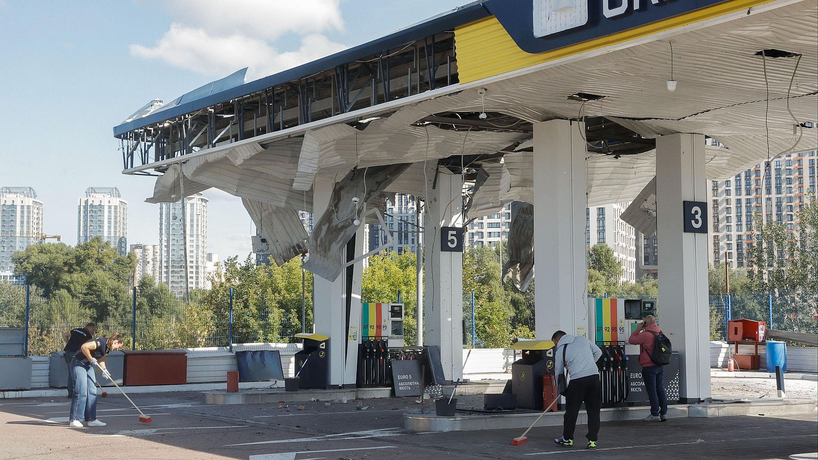 Gasolinera en Kiev, Ucrania, dañada por un ataque ruso el 15/09/2026. Trabajadores limpian escombros bajo un cielo nublado, con edificios altos al fondo.