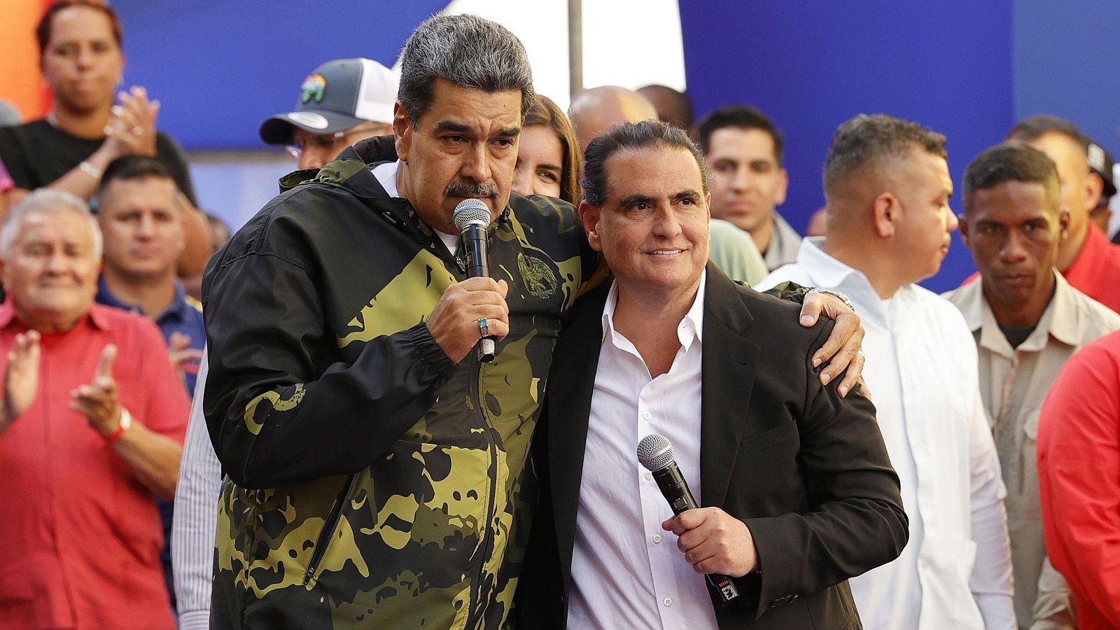Nicolás Maduro y Alex Saab en un evento público al aire libre. Maduro viste chaqueta de camuflaje y Saab traje oscuro. Ambos sostienen micrófonos.