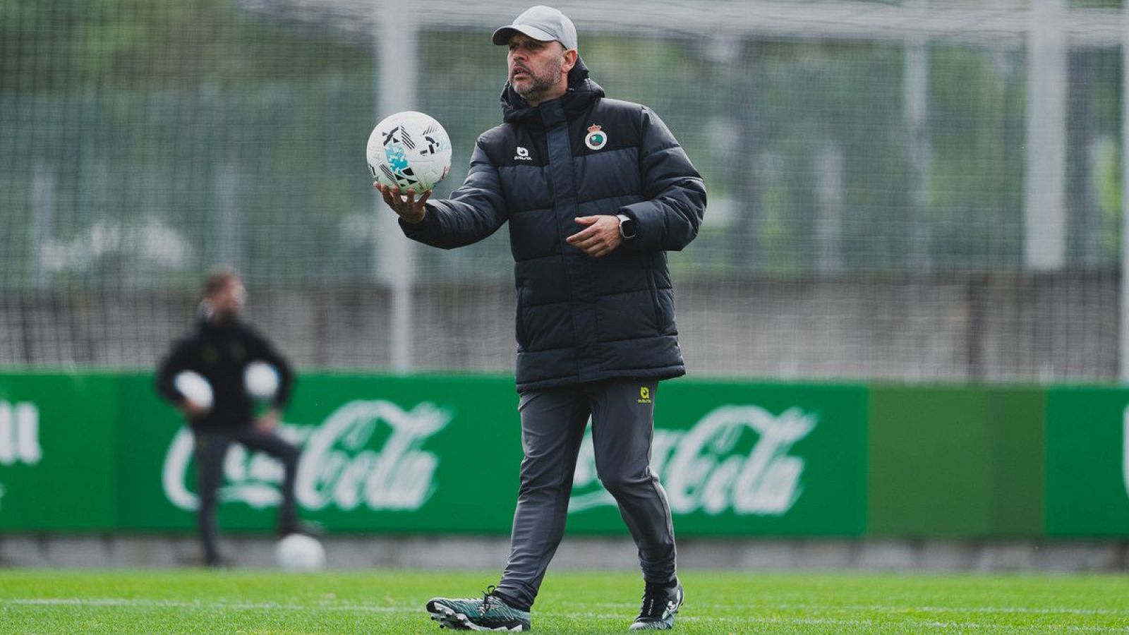Entrenador del Racing, José Alberto López, con balón en mano, viste ropa deportiva gris y chaqueta oscura. Fondo con muro y logo de Coca-Cola.