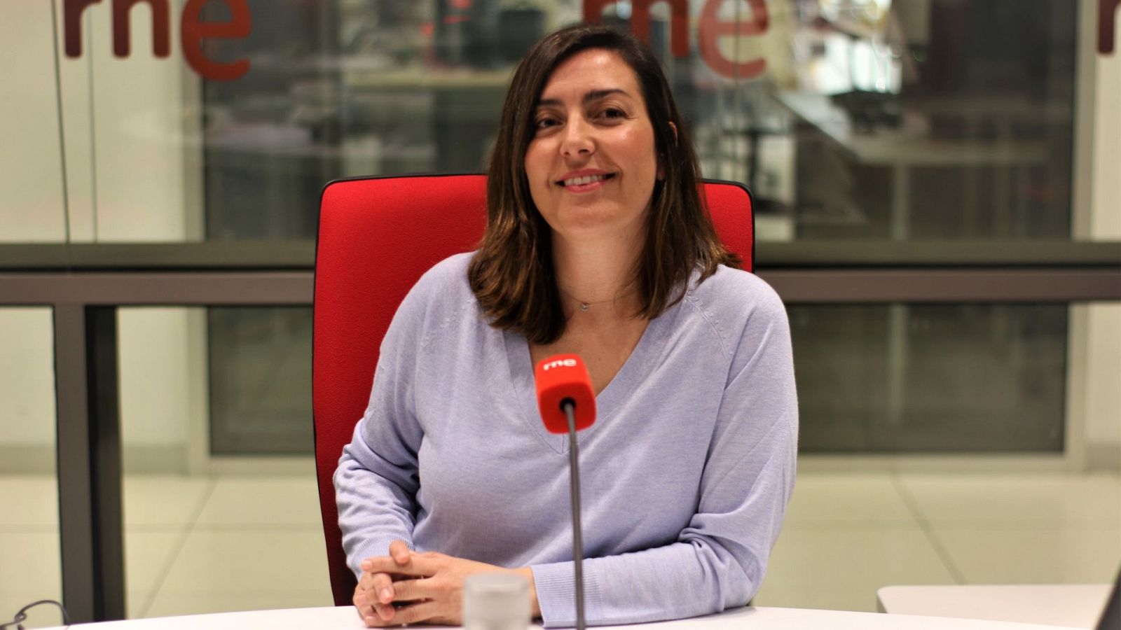 Laura Barrachina, periodista cultural de RTVE, en un estudio de radio con el logo "rne" visible. La imagen la muestra con una blusa lila y una expresión sonriente.