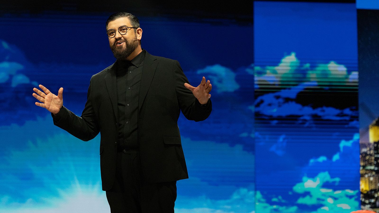 Manu Sánchez, presentador, gesticula en un escenario con traje negro y gafas, frente a pantallas con cielo y ciudad.