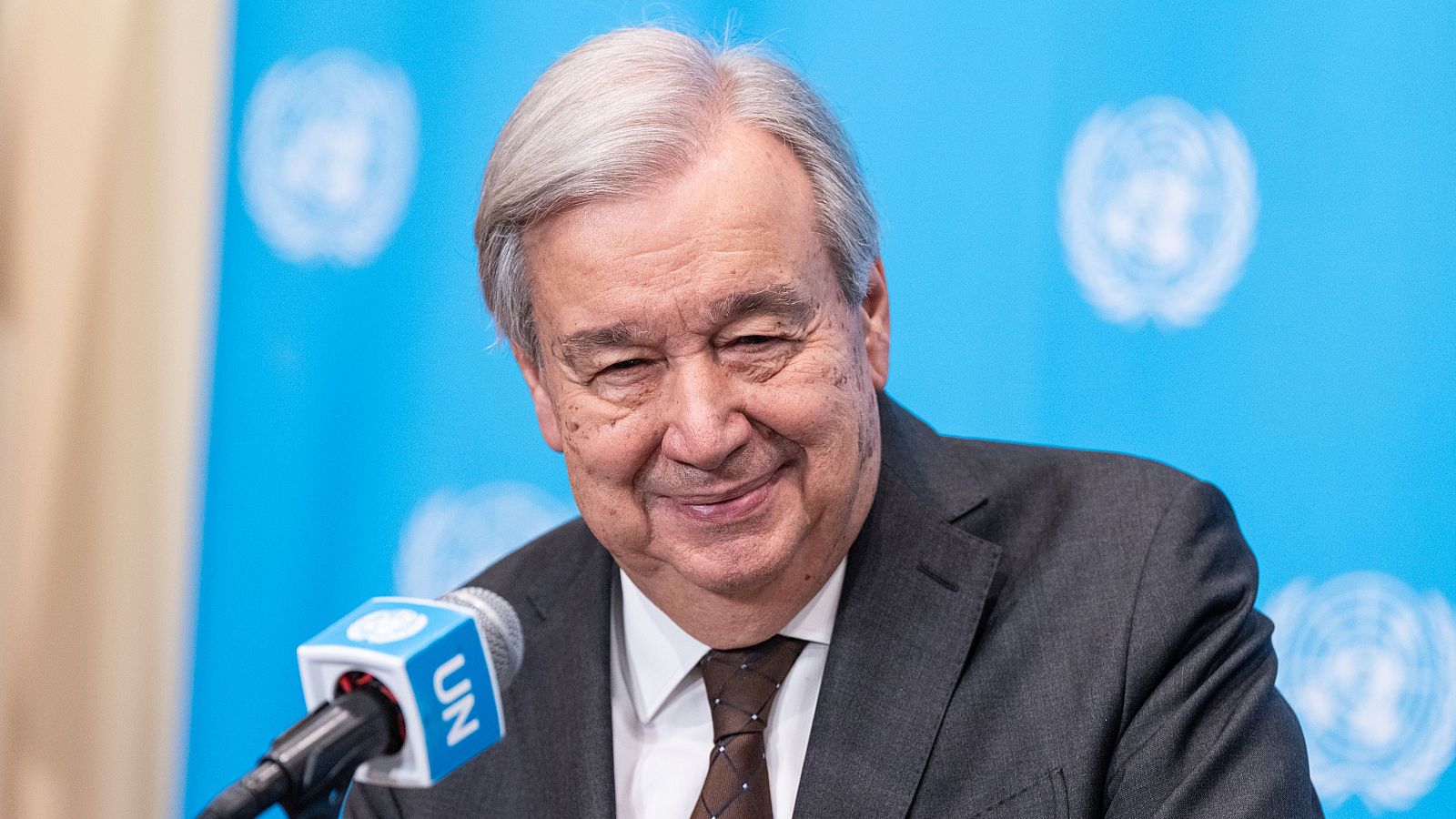António Guterres, Secretario General de la ONU, sonríe con un traje gris y corbata marrón, con un micrófono de la ONU a su lado y un fondo azul claro con logotipos de la organización.
