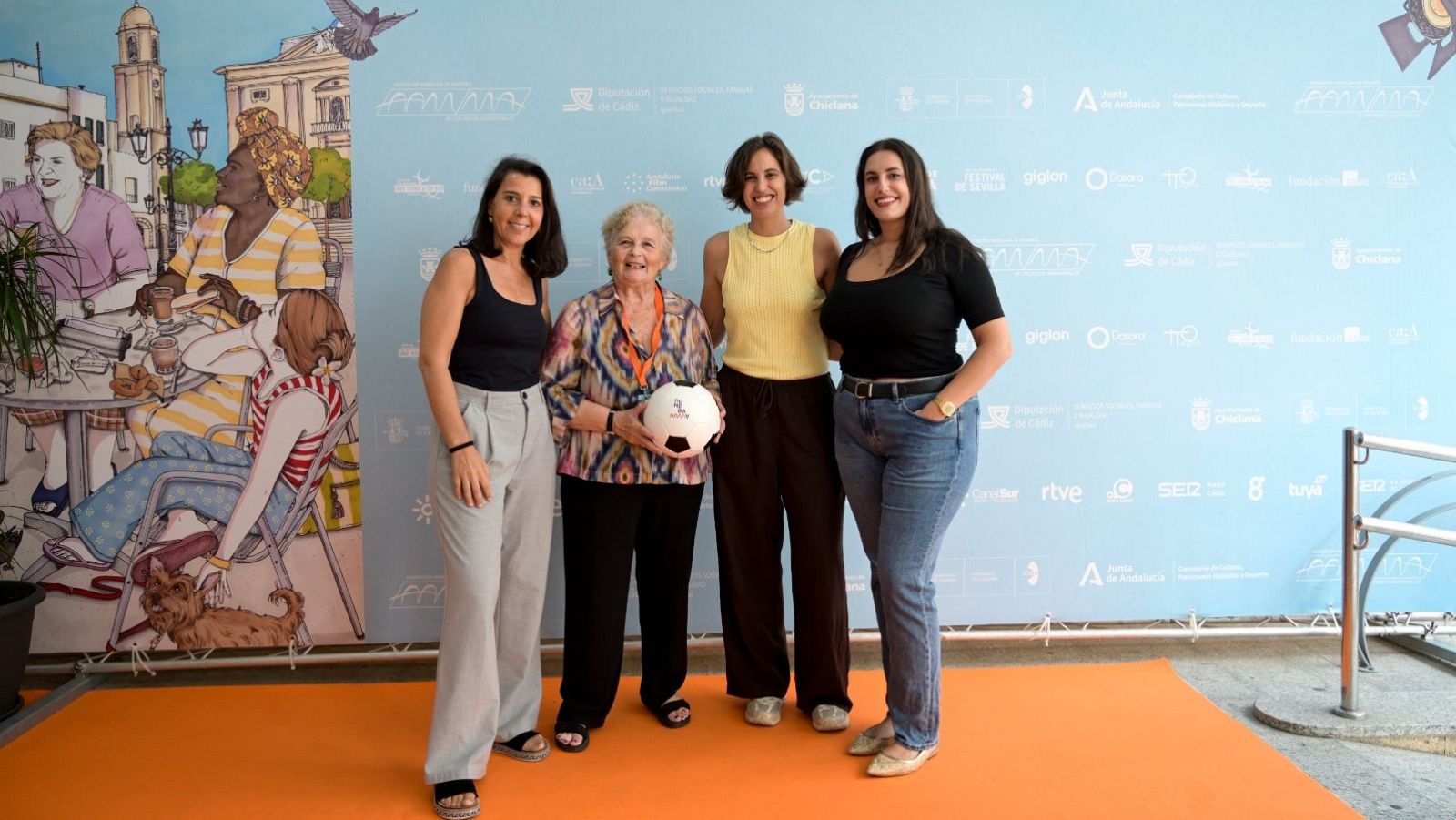 Pilar Crespo, Carmen Arce (con un balón de fútbol), Marta Díaz y Beatriz Aparicio posan sonrientes en un evento con un fondo de mural y logotipos de medios.