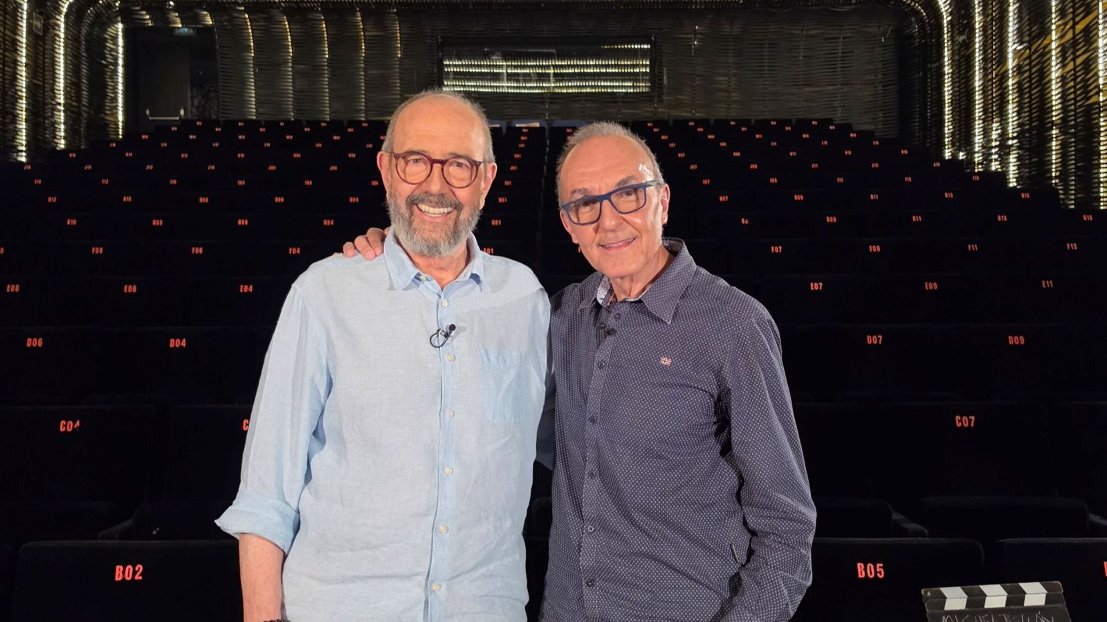 Miguel Rellán y Gerardo Sánchez sonríen en un auditorio. Rellán lleva gafas y camisa azul claro con micrófono. Sánchez, con gafas azules y camisa oscura, apoya su brazo en Rellán.