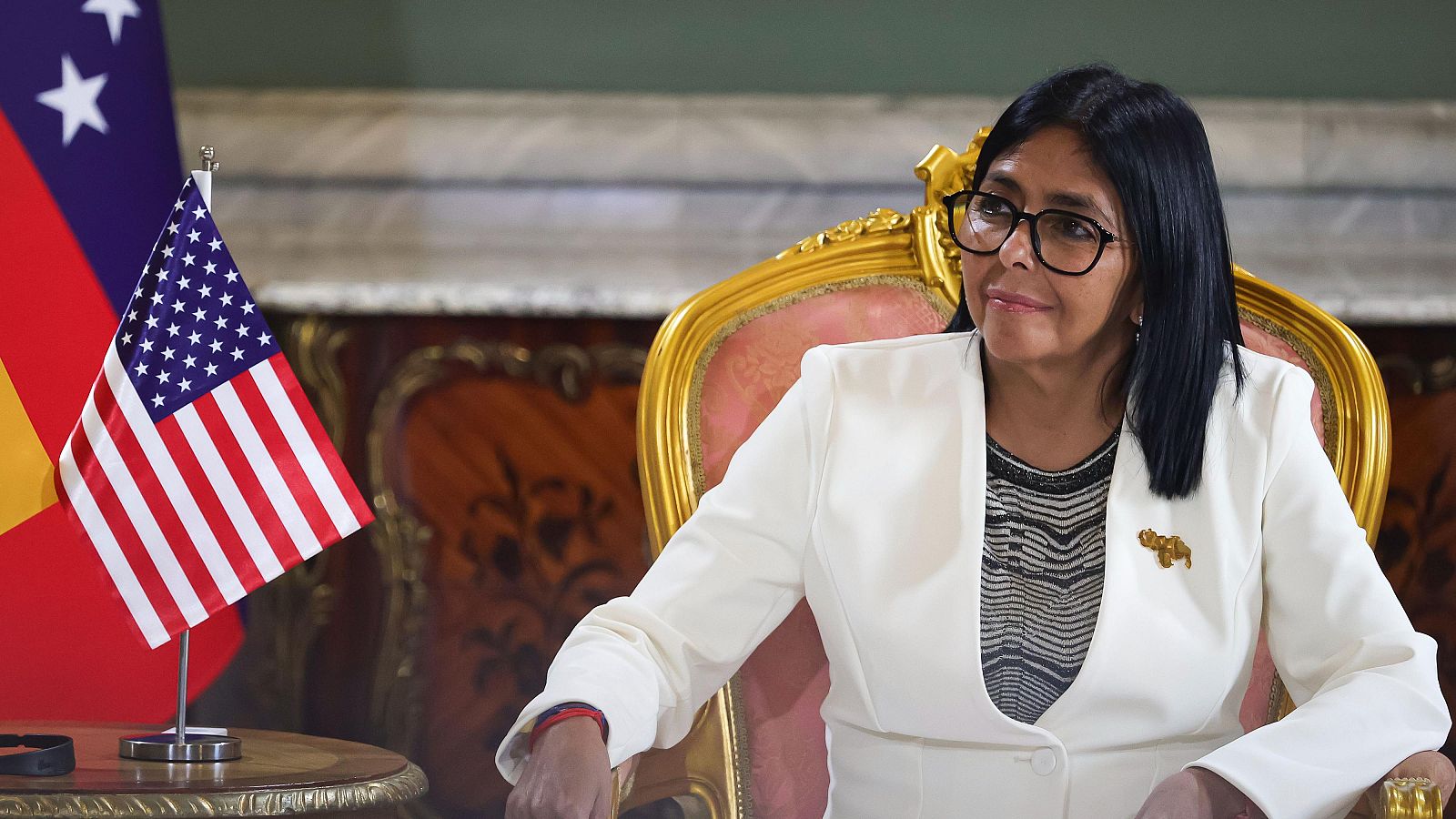 Delcy Rodríguez, presidenta interina de Venezuela, con blazer blanco y broche dorado, sentada en sillón clásico. Banderas de EE.UU. y Venezuela presentes. Rueda de prensa diplomática.