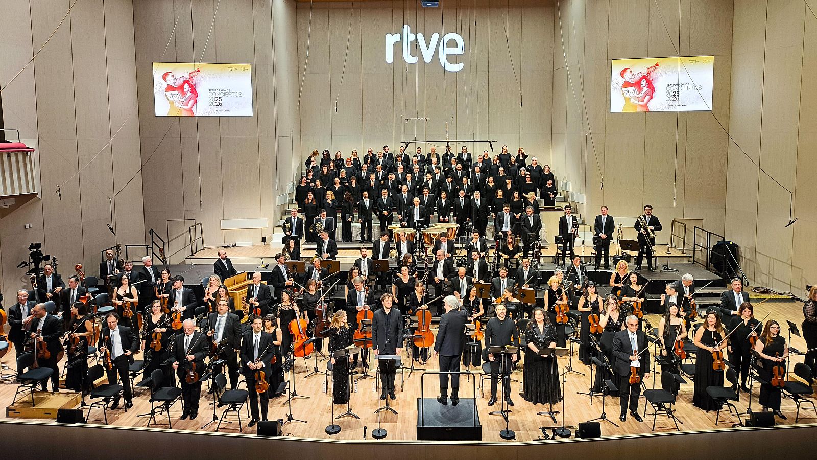 Vista panorámica de un concierto de la Orquesta y Coro RTVE, con músicos y cantantes en el escenario, un logo de "rtve" y pantallas promocionales.