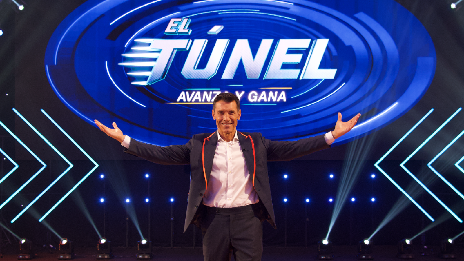 Jesús Vázquez presenta "EL TÚNEL" con el lema "AVANZA Y GANA" en un escenario iluminado con luces azules y detalles naranjas en su traje.