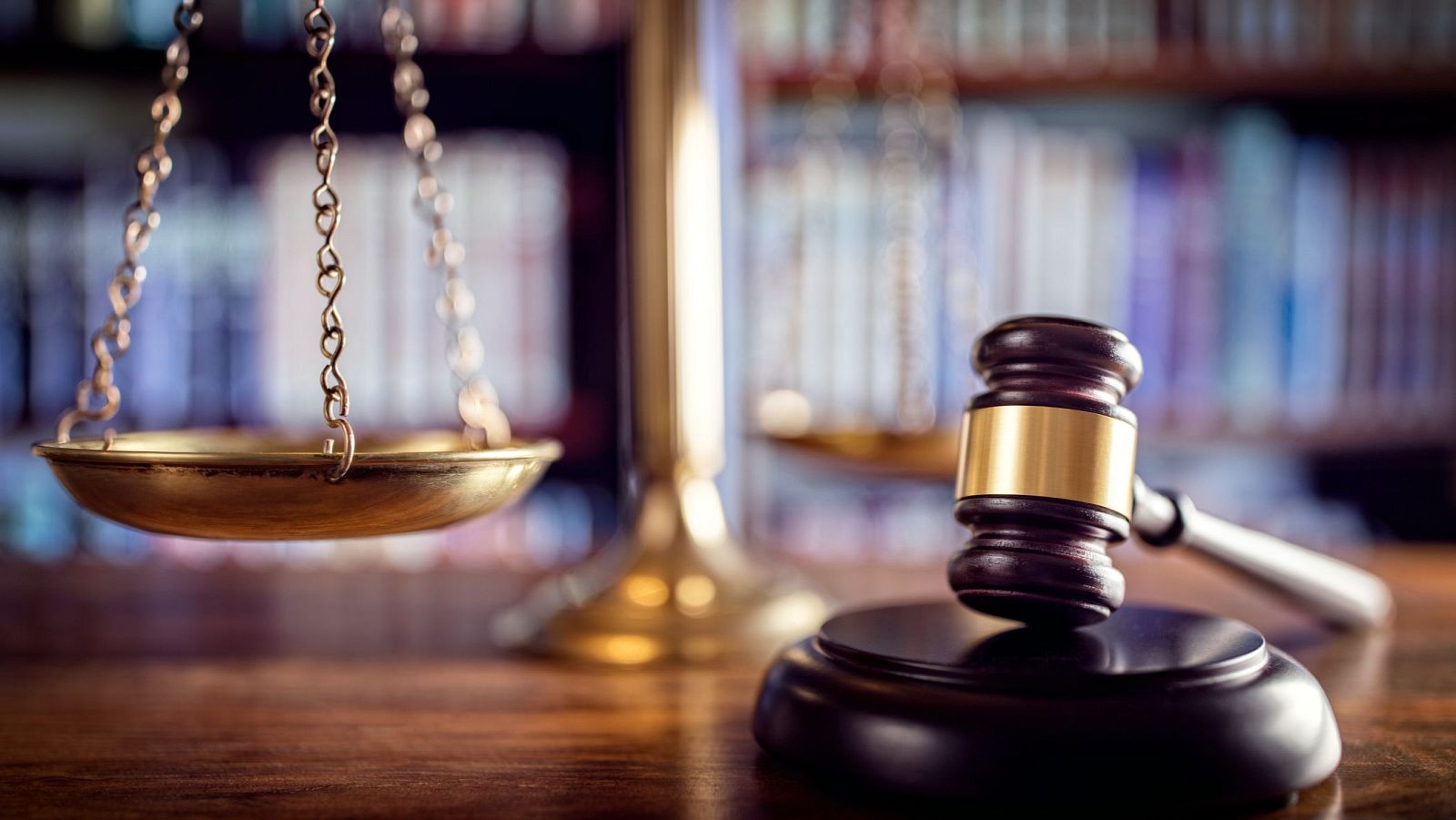 Martillo de juez y balanza de la justicia sobre superficie de madera pulida, con fondo de estanterías de libros desenfocadas. Símbolos del sistema judicial.