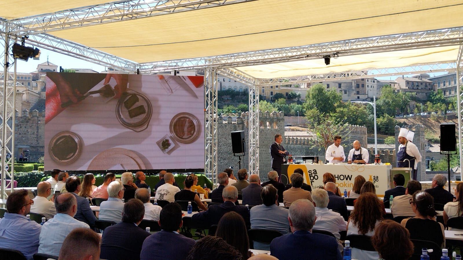 Iván Cerdeño, chef con chaquetilla blanca y delantal azul, dirige una demostración culinaria en Toledo. Una pantalla LED muestra el proceso de cocina ante una audiencia.