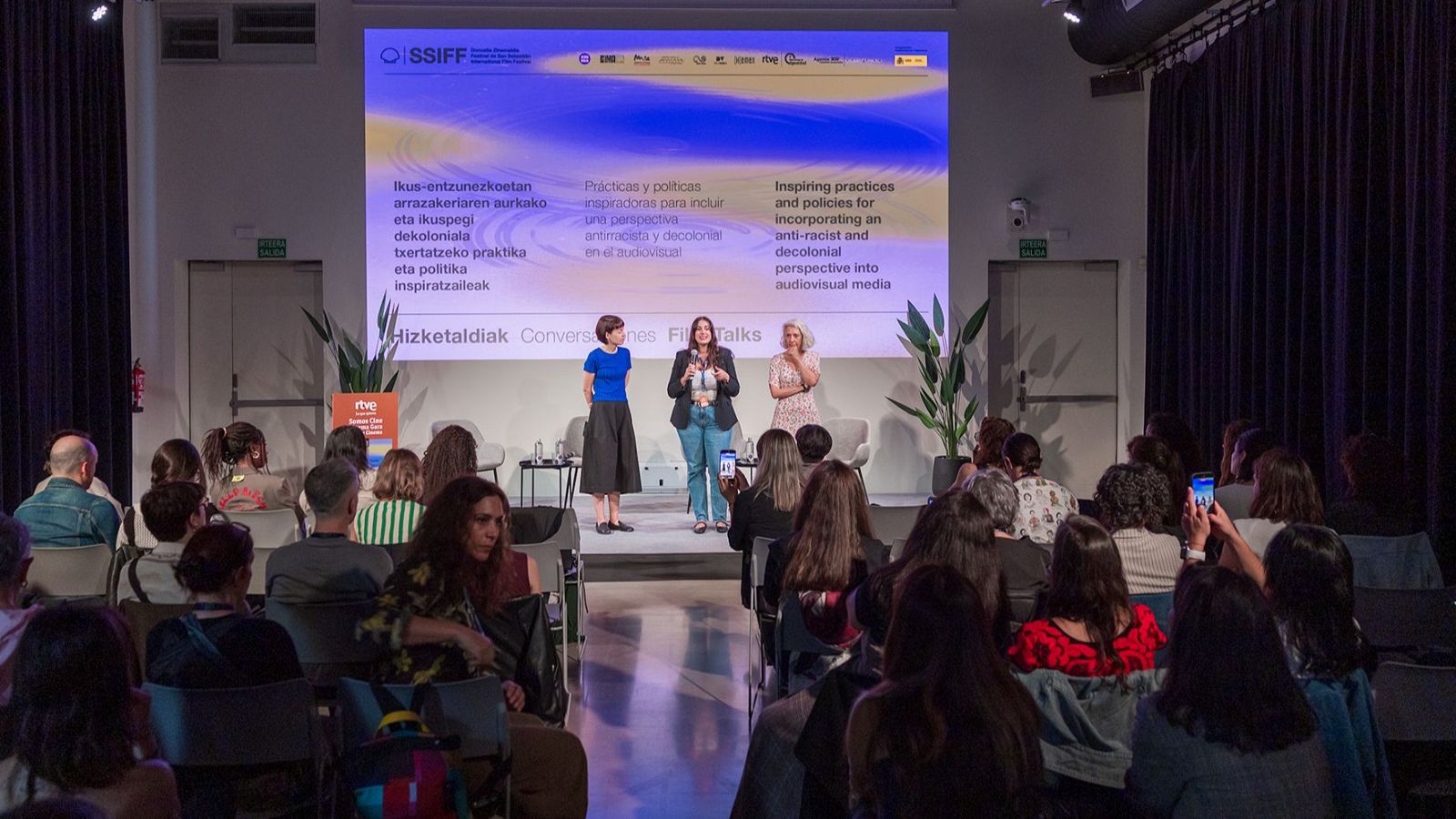 María Eizaguirre de RTVE, flanqueada por dos ponentes, presenta en el SSIFF sobre antirracismo y decolonialismo en el audiovisual, con el logo de RTVE y el del festival visibles.