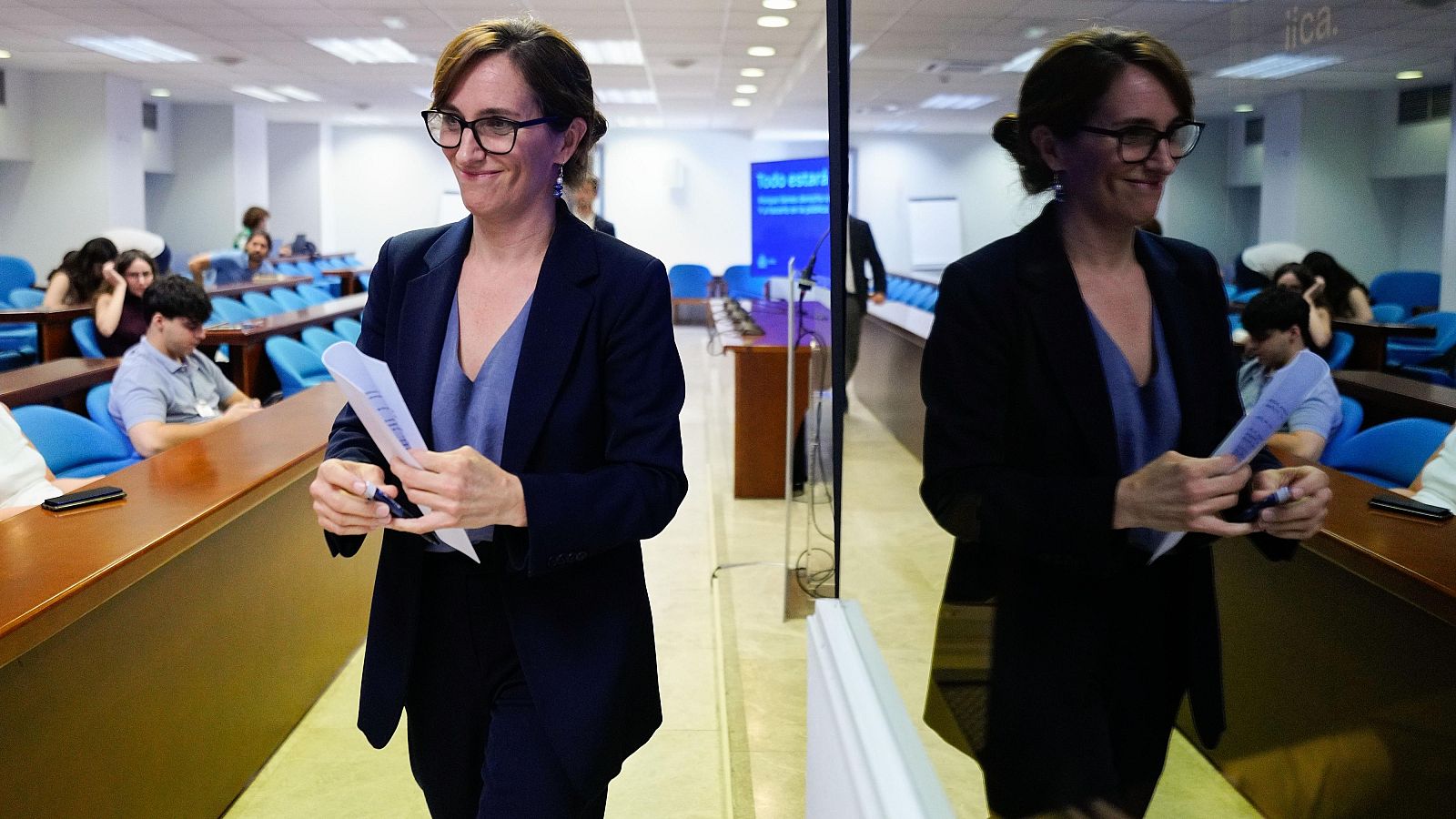Mónica García, Ministra de Sanidad, sonríe mientras camina con papeles y bolígrafo, reflejada en cristal. Al fondo, público y pantalla azul con texto parcial.
