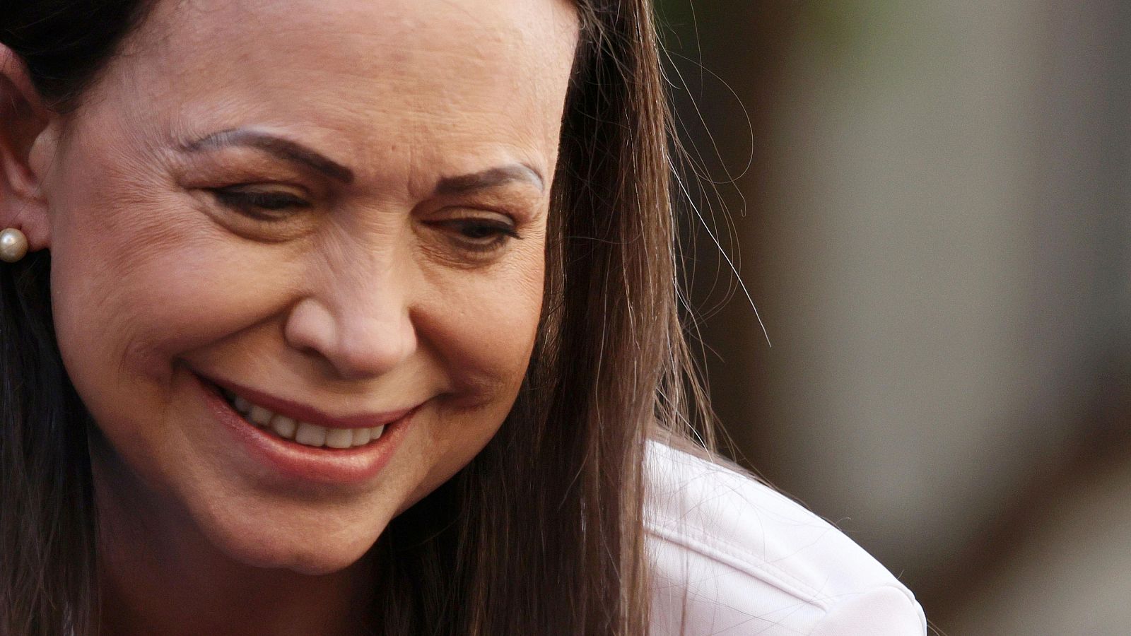 Retrato de Maria Corina Machado, política venezolana, con cabello castaño oscuro, camisa blanca y pendiente de perla, sonriendo suavemente.