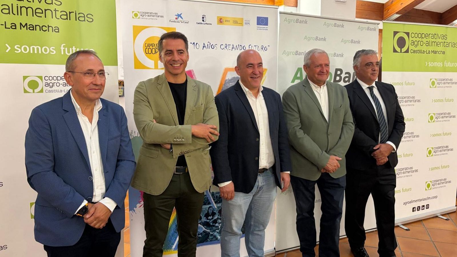 Cinco hombres posan en fila frente a paneles informativos de "Cooperativas Agro-alimentarias Castilla-La Mancha" y "CAMPUS COOP" en un evento.