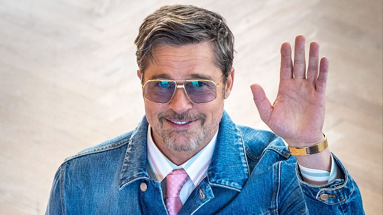 Primer plano de Brad Pitt saludando con la mano, sonriendo y luciendo gafas de sol aviador y chaqueta vaquera. Fondo blanco desenfocado.