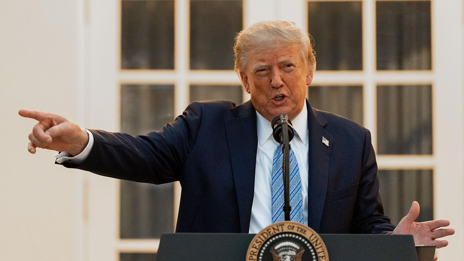 Donald Trump, con traje azul y pin de EE.UU., habla enérgicamente desde un podio con el sello presidencial visible. Su brazo izquierdo apunta y la mano derecha está levantada.