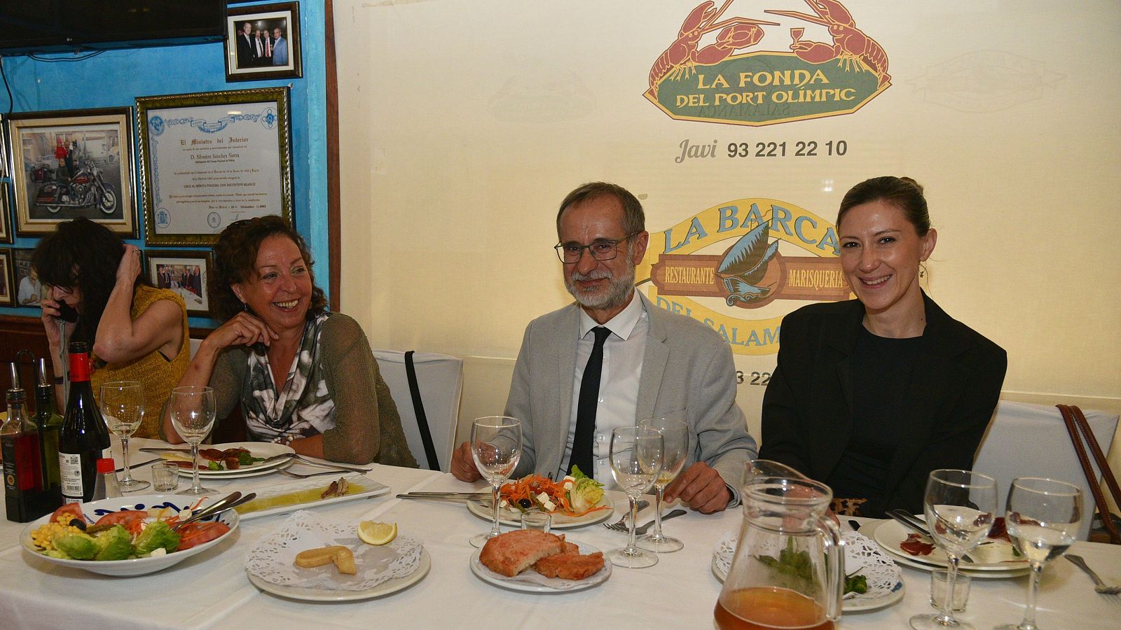 Miquel Iceta y Eva Granados en un restaurante, rodeados de comida y bebida. Iceta viste traje gris y gafas, Granados chaqueta negra. Ambos sonríen a la cámara.