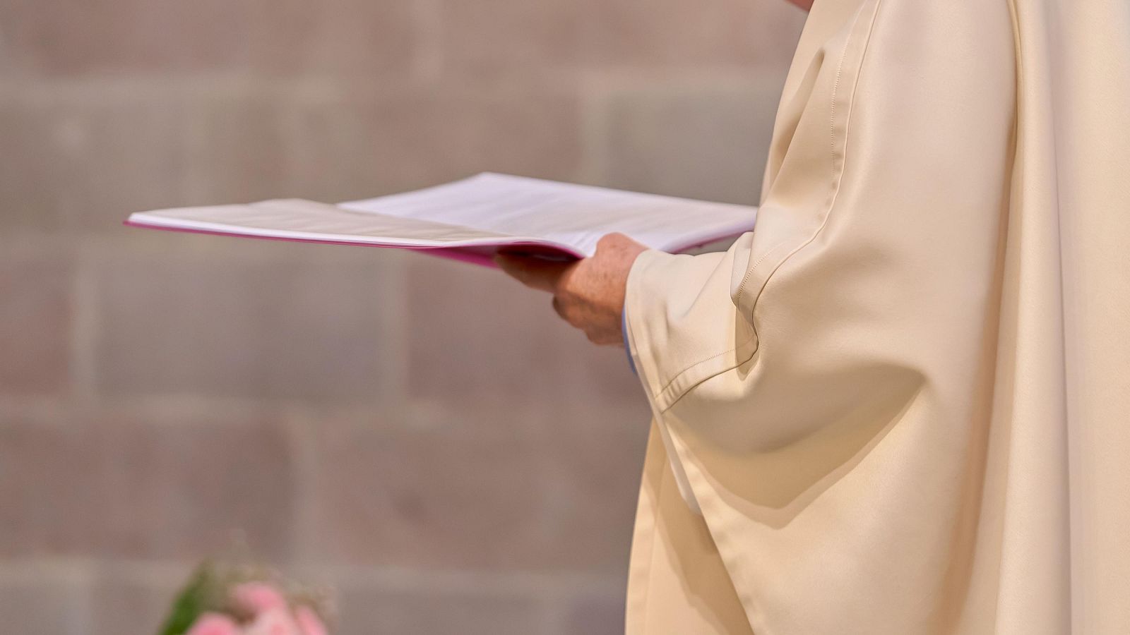 Sacerdote con túnica crema y libro de cubierta rosa fucsia, en interior de piedra. Fondo borroso verde y rosa.