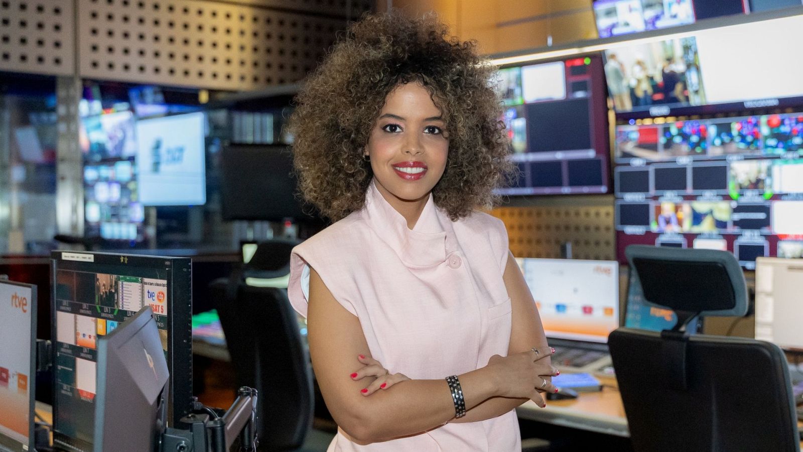 Periodista Ebbaba Hameida sonríe a cámara en una sala de control de RTVE, vestida con blusa rosa y accesorios. El fondo muestra monitores y pantallas.
