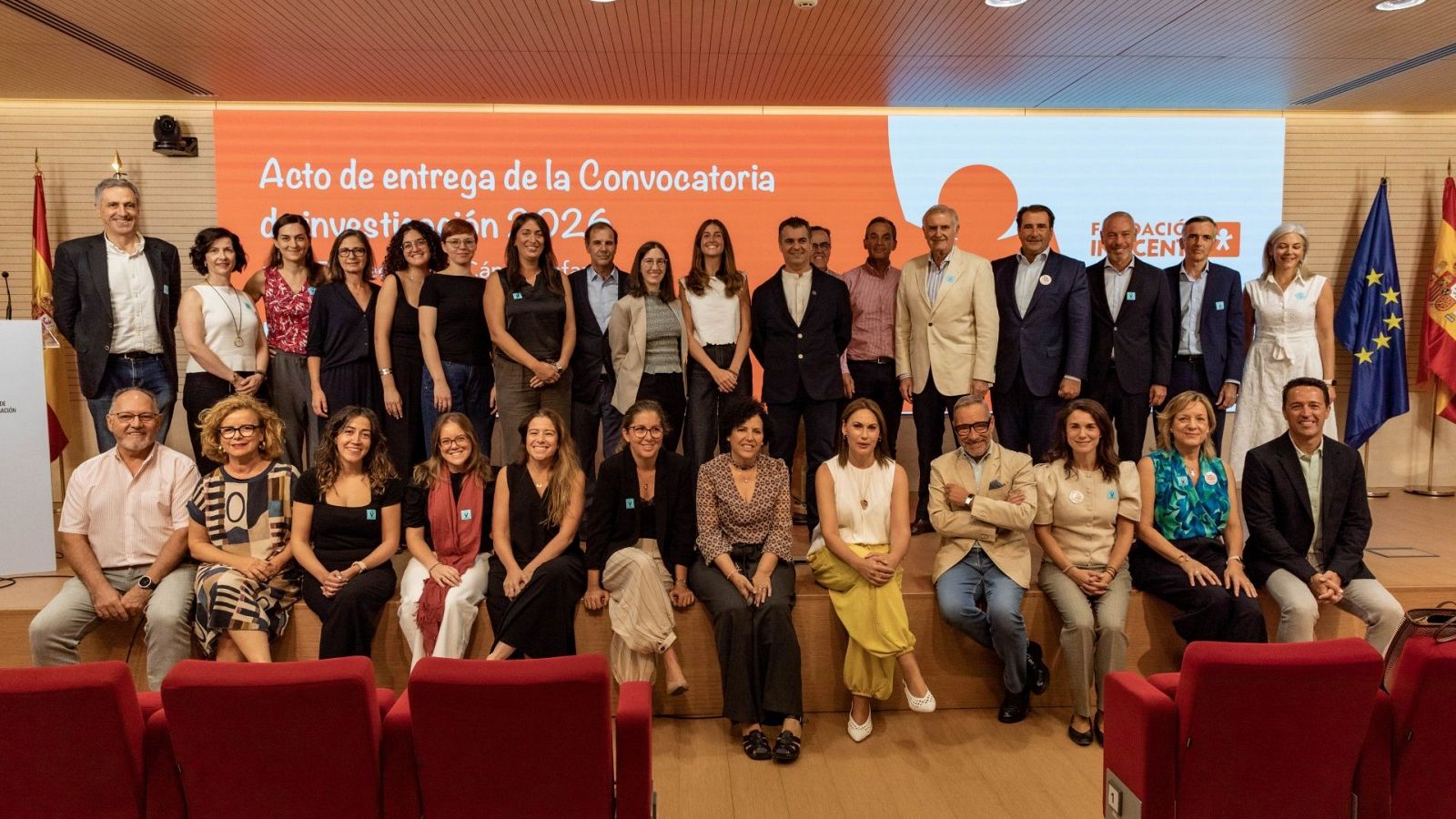 Fotografía de grupo en el "Acto de entrega de la Convocatoria de Investigación 2026" de la Fundación Inocente, con Juan y Medio presente. Se observan dos filas de asistentes, una pantalla con el título del evento y banderas institucionales.