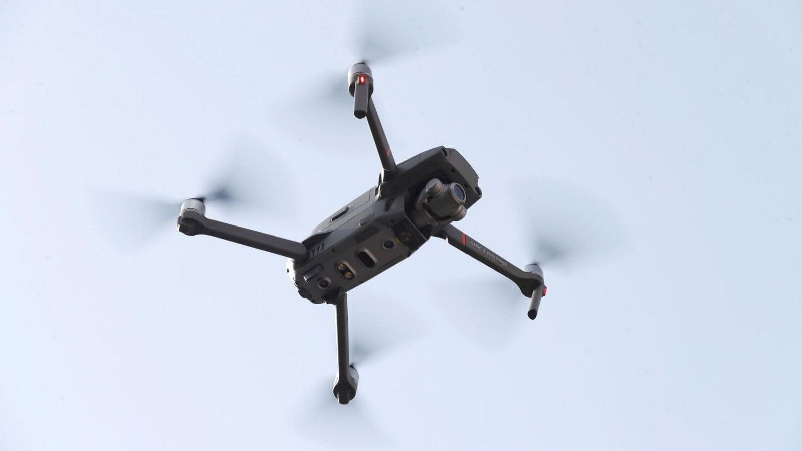 Dron cuadricóptero oscuro en vuelo, con cámara frontal y hélices en movimiento, bajo un cielo azul pálido. Luces rojas visibles en los brazos.