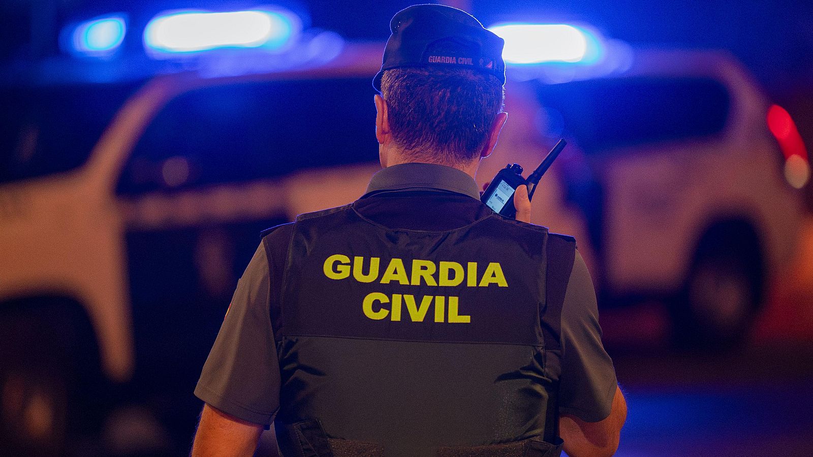 Agente de la Guardia Civil de espaldas, escuchando por walkie-talkie, con uniforme y chaleco "GUARDIA CIVIL" amarillo. Vehículo patrulla con luces azules al fondo.