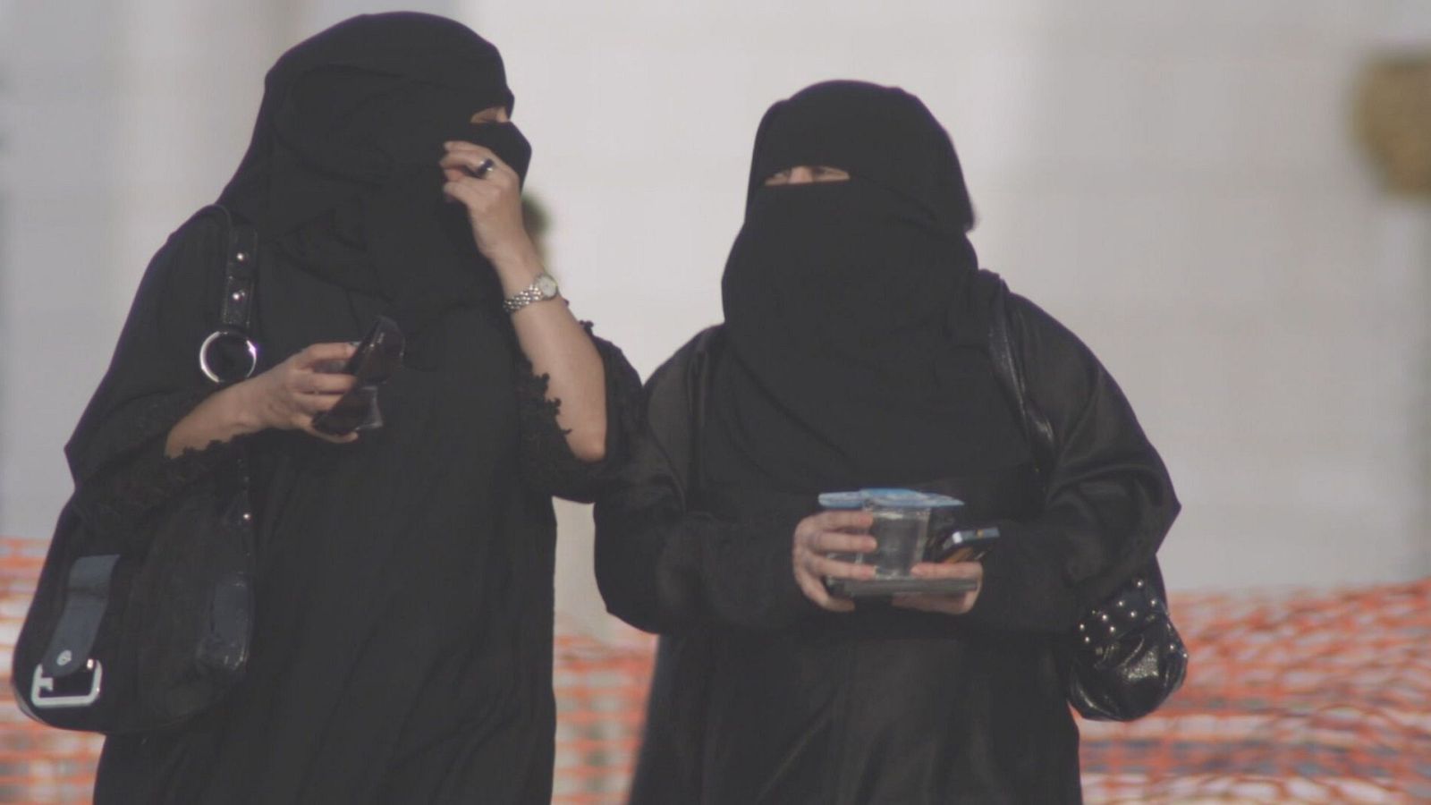 Dues figures amb vestimenta tradicional negra, niqabs i hijabs, es troben una al costat de l'altra. Una porta rellotge i anell, l'altra subjecta un recipient blau.